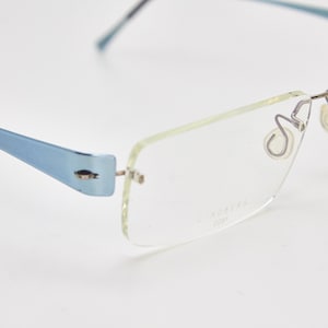 May include: A pair of light blue rimless spectacles with clear lenses. The glasses have a silver metal frame and a small silver metal detail on the side of the frame. The text "CK8244 2001" is printed on the right lens.