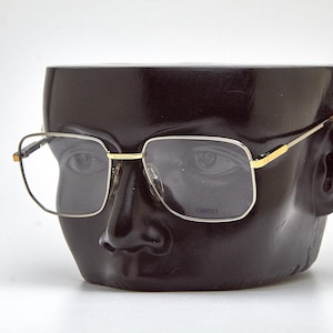 Puede incluir: Gafas con montura dorada y lentes cuadradas sobre una cabeza de maniquí negra. Las gafas tienen un puente y patillas doradas, con un diseño sutil en las patillas. La cabeza del maniquí es negra brillante.