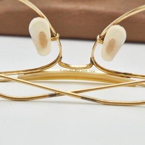 May include: Gold-tone metal eyeglasses featuring a thin wire frame, cream-colored nose pads, and a bridge engraved with the text 1/20 10K GF USA. The temples are folded inward, showcasing the metallic construction and the detailed bridge markings.