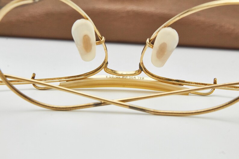 May include: Gold-tone metal eyeglasses featuring a thin wire frame, cream-colored nose pads, and a bridge engraved with the text 1/20 10K GF USA. The temples are folded inward, showcasing the metallic construction and the detailed bridge markings.