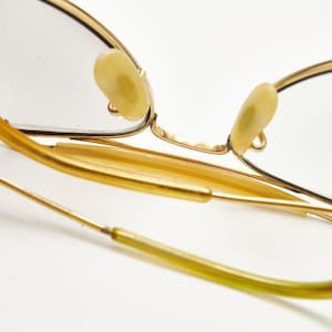 Puede incluir: Primer plano de gafas con montura dorada de dise&ntilde;o cl&aacute;sico. Las gafas presentan una montura fina y delicada y almohadillas nasales de color claro. Las patillas son de color oro c&aacute;lido y las lentes son transparentes. La est&eacute;tica general es vintage y elegante.