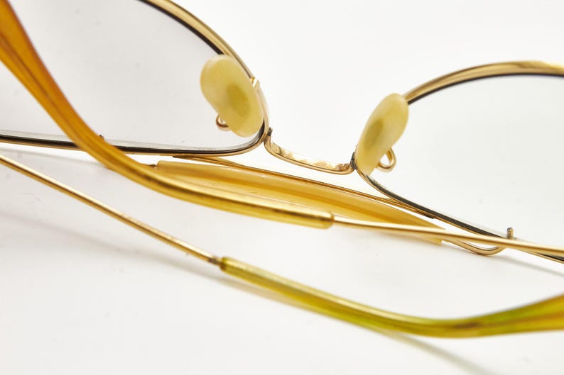 Puede incluir: Primer plano de gafas con montura dorada de dise&ntilde;o cl&aacute;sico. Las gafas presentan una montura fina y delicada y almohadillas nasales de color claro. Las patillas son de color oro c&aacute;lido y las lentes son transparentes. La est&eacute;tica general es vintage y elegante.
