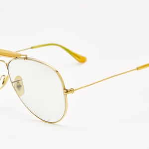 Puede incluir: Gafas de aviador con montura dorada, lentes transparentes y puente dorado. Las patillas son doradas con puntas amarillas. Las lentes son de color gris claro. Las gafas est&aacute;n sobre un fondo blanco.