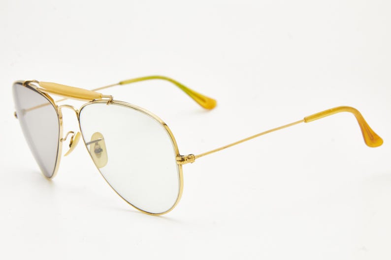 Puede incluir: Gafas de aviador con montura dorada, lentes transparentes y puente dorado. Las patillas son doradas con puntas amarillas. Las lentes son de color gris claro. Las gafas est&aacute;n sobre un fondo blanco.