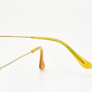 Puede incluir: Primer plano de gafas de sol aviador con montura dorada y patilla verde claro y amarilla. El puente nasal y la montura son dorados. Las lentes son de color gris claro. Las gafas de sol est&aacute;n sobre un fondo blanco.
