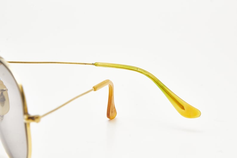 Puede incluir: Primer plano de gafas de sol aviador con montura dorada y patilla verde claro y amarilla. El puente nasal y la montura son dorados. Las lentes son de color gris claro. Las gafas de sol est&aacute;n sobre un fondo blanco.