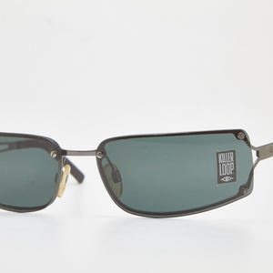 Op de afbeelding: Donkergrijze zonnebril met rechthoekige groene glazen. De zonnebril heeft een dun, donkergrijs montuur en de tekst "KILLER LOOP" is zichtbaar op het rechterglas. De pootjes zijn donkerder grijs.