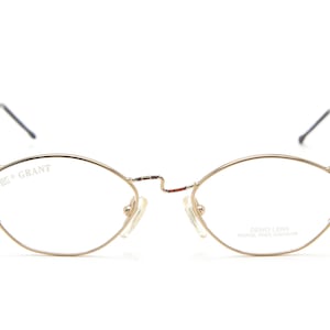 May include: Gold-toned metal spectacles featuring thin, oval-shaped frames with a bridge detail. The left lens is etched with the text GRANT, while the right lens displays the text DEMO LENS WORLD FREE DIVISION. The temples are thin and metallic.