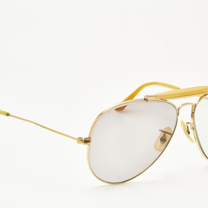 Puede incluir: Gafas de sol de aviador con montura dorada y lentes claras. Las gafas tienen un doble puente y una barra en la frente. Las patillas son doradas con puntas amarillas. Las lentes son de color gris claro.