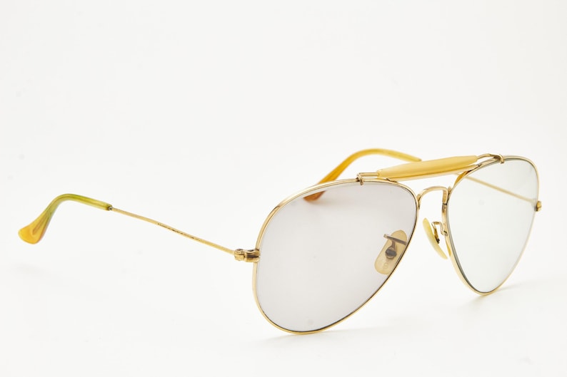 Puede incluir: Gafas de sol de aviador con montura dorada y lentes claras. Las gafas tienen un doble puente y una barra en la frente. Las patillas son doradas con puntas amarillas. Las lentes son de color gris claro.