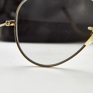 Puede incluir: Primer plano de gafas con montura dorada, montura negra y lentes transparentes. Las gafas tienen un estilo aviador cl&aacute;sico, con una fina montura de alambre dorado y almohadillas nasales. El fondo es una forma negra borrosa.