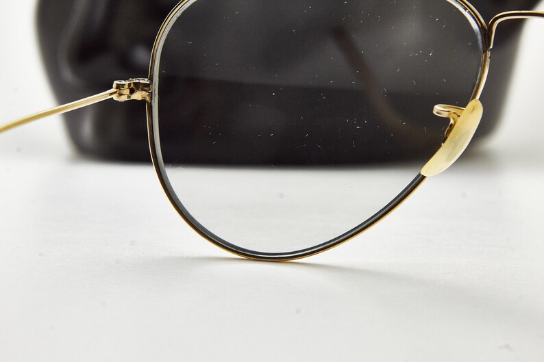 Puede incluir: Primer plano de gafas con montura dorada, montura negra y lentes transparentes. Las gafas tienen un estilo aviador cl&aacute;sico, con una fina montura de alambre dorado y almohadillas nasales. El fondo es una forma negra borrosa.