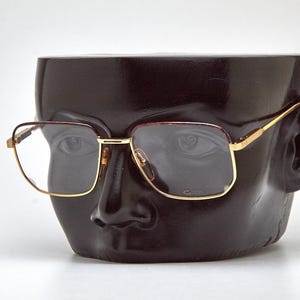 May include: Gold-rimmed eyeglasses with a rectangular lens shape. The glasses have a brown and gold color scheme and are displayed on a black mannequin head. The glasses are a vintage style.