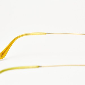 Puede incluir: Primer plano de unas gafas de sol de aviador con montura dorada. Las patillas son de color amarillo transl&uacute;cido y la montura es de oro fino y brillante. Las lentes no son visibles. Las gafas est&aacute;n sobre un fondo blanco.