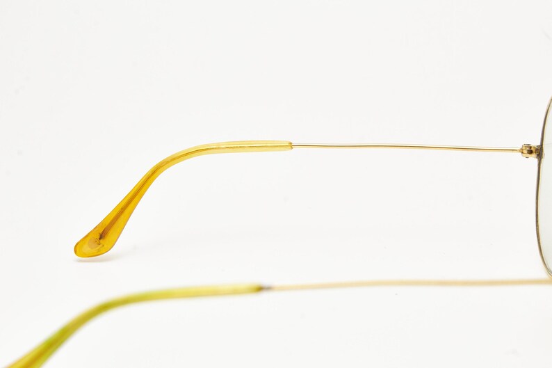 Puede incluir: Primer plano de unas gafas de sol de aviador con montura dorada. Las patillas son de color amarillo transl&uacute;cido y la montura es de oro fino y brillante. Las lentes no son visibles. Las gafas est&aacute;n sobre un fondo blanco.