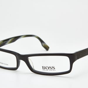 Könnte beinhalten: Schwarze rechteckige Brille mit klaren Gläsern. Die Fassungen haben eine glänzende Oberfläche und die Bügel weisen ein grün-braunes Muster auf. Der Markenname "BOSS" ist auf dem Glas abgebildet.