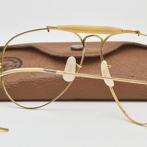 May include: Gold-tone wire-frame aviator glasses featuring a cream-colored brow bar, curved cable temples, and clear nose pads. The glasses are positioned in front of a brown textured leather-like case with a visible circular logo stamp.