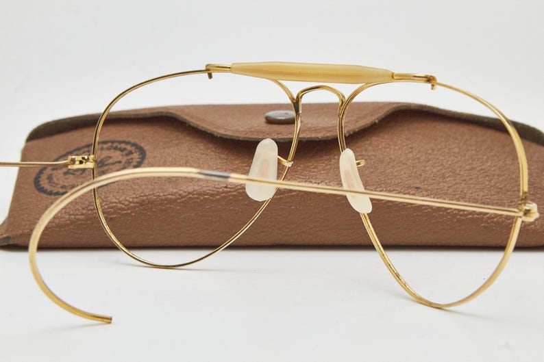 May include: Gold-tone wire-frame aviator glasses featuring a cream-colored brow bar, curved cable temples, and clear nose pads. The glasses are positioned in front of a brown textured leather-like case with a visible circular logo stamp.