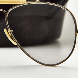 Puede incluir: Primer plano de gafas de sol de aviador con montura dorada y lentes grises. Las gafas de sol tienen una forma cl&aacute;sica de l&aacute;grima y una montura y patillas doradas. Las almohadillas nasales son de color beige claro. El fondo es una forma negra borrosa.