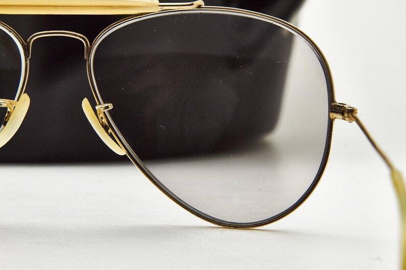 Puede incluir: Primer plano de gafas de sol de aviador con montura dorada y lentes grises. Las gafas de sol tienen una forma cl&aacute;sica de l&aacute;grima y una montura y patillas doradas. Las almohadillas nasales son de color beige claro. El fondo es una forma negra borrosa.