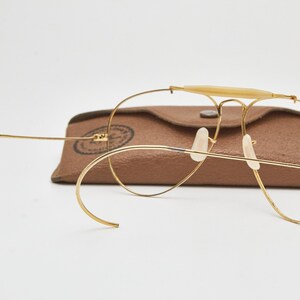 May include: Gold-tone wire-frame aviator glasses featuring a brow bar with a cream-colored insert, curved cable temples, and clear nose pads. The glasses are positioned in front of a brown textured leather-like protective carrying case.