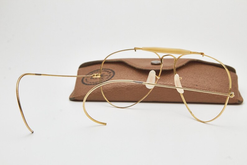 May include: Gold-tone wire-frame aviator glasses featuring a brow bar with a cream-colored insert, curved cable temples, and clear nose pads. The glasses are positioned in front of a brown textured leather-like protective carrying case.