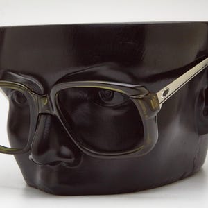 May include: Dark olive green eyeglasses with a square frame design. The glasses have silver-toned temples and are displayed on a black mannequin head. The overall style is retro and classic.