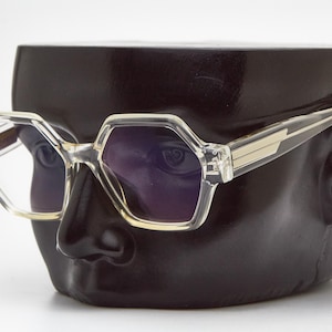 May include: Clear frame sunglasses with a geometric shape and dark lenses. The sunglasses are on a black mannequin head.
