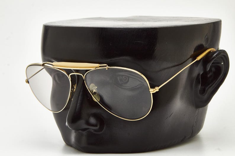 Puede incluir: Gafas de sol de aviador con montura dorada y lentes oscuras. Las gafas de sol tienen un doble puente y una barra de color claro sobre el puente nasal. Las gafas se muestran en una cabeza de maniqu&iacute; negra.