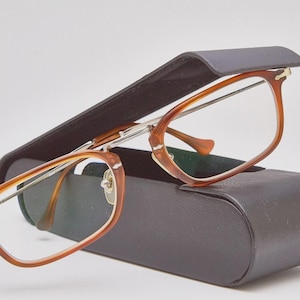 Puede incluir: Un par de gafas con montura marrón y lentes transparentes descansa sobre un estuche para gafas marrón oscuro. Las gafas tienen un diseño clásico con forma rectangular. El estuche es rectangular y tiene una parte superior curvada.