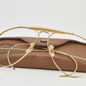 May include: Gold-tone metal aviator-style eyeglasses with a thin wire frame and a brow bar, positioned in front of a brown textured leather-like protective case with a snap button closure and a visible circular logo stamp on the left side.