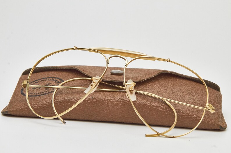 May include: Gold-tone metal aviator-style eyeglasses with a thin wire frame and a brow bar, positioned in front of a brown textured leather-like protective case with a snap button closure and a visible circular logo stamp on the left side.