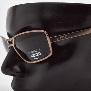 May include: Brown and gold-toned sunglasses with dark lenses. The sunglasses have the word "KENZO" printed on the side.