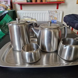 May include: A stainless steel tea set on a silver tray. The set includes a teapot, a creamer, a sugar bowl, and a tall pitcher. The items are arranged on a rectangular tray. The background is a dark table.