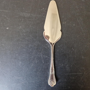 Puede incluir: Servidor de pastel plateado con un mango decorativo y un borde festoneado. El servidor está hecho de un metal brillante y reflectante y está diseñado para servir pastel o tarta. El mango presenta un diseño ornamentado.