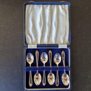 May include: A set of six silver spoons in a blue velvet-lined case. The spoons have a classic design with rounded bowls and slender handles. The case is open, revealing the spoons nestled in individual slots.