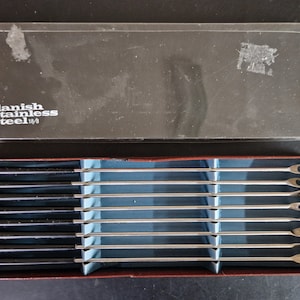 May include: A set of eight silver cocktail sticks with black handles in a red and blue box. The box has the text "Danish Stainless Steel 1/8" printed on it.