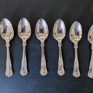 Op de afbeelding: Zes zilveren lepels in een rij gerangschikt. Elke lepel heeft een afgeronde kom en een decoratief handvat met een schelpachtig ontwerp. De lepels zijn zeer reflecterend en tonen de omgeving. De lepels liggen op een donkere ondergrond.