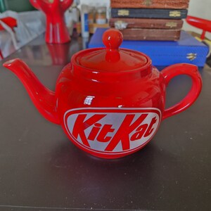 May include: A red ceramic teapot with a white "Kit Kat" logo. The teapot has a rounded body and a spout on the left side.