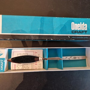 May include: An open box containing an Oneida Craft solid stainless steel palette knife. The knife has a dark handle and a silver blade with a floral design. The box is teal and white with the brand name "Oneida CRAFT" printed on it.
