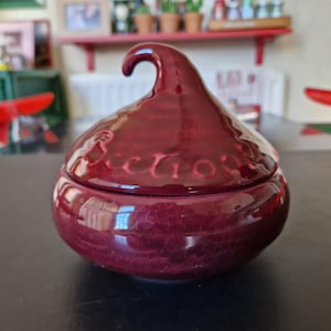 May include: A maroon ceramic candy dish with a unique, rounded shape and a curved handle on the lid. The dish has a glossy finish and the word "Kisses" is embossed around the lid. The dish is sitting on a dark surface.