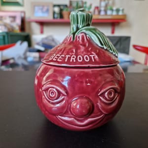 May include: A ceramic beet-shaped jar with a lid, painted in a deep red hue. The jar features a face with eyes, a nose, and a smiling mouth. The lid is green and has the word "BEETROOT" in white.