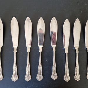Op de afbeelding: Een set van acht zilveren botermessen, gerangschikt op een donkere ondergrond. Elk mes heeft een breed, afgerond lemmet en een decoratief handvat met een sierlijk, schelpachtig ontwerp. De messen reflecteren het licht.