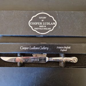 May include: A silver knife with a decorative handle, presented in a black box. The box lid displays "CUTLERY BY COOPER LUDLAM MADE IN SHEFFIELD ENGLAND". The knife is in a black velvet-lined compartment.