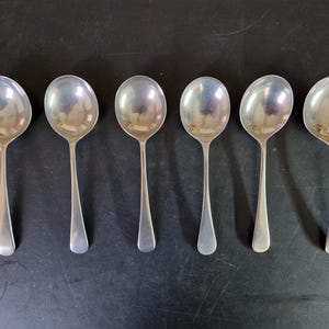 May include: Six silver soup spoons displayed in a line. Each spoon features a rounded bowl and a long, thin handle. The spoons are made of a shiny, reflective metal, possibly stainless steel or silver, set against a dark surface.