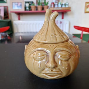 May include: A tan ceramic onion-shaped container with a lid. The container has a face with closed eyes and tear drops. The lid is shaped like the top of an onion and has the word "Onions" on it. The container is on a black surface.