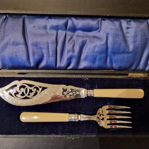 Puede incluir: Un juego de servir pescado antiguo, que incluye un cuchillo y un tenedor, presentado en una caja negra con un forro de terciopelo azul. El cuchillo presenta una hoja plateada ornamentada con un diseño decorativo, y ambos utensilios tienen mangos color crema.