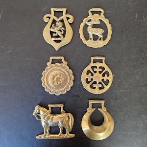 Peut inclure: Un ensemble de six ornements de harnais en laiton représentant divers motifs dont un lion, un cerf, une étoile, un motif géométrique, un cheval et une forme de croissant. Chaque pièce possède une boucle supérieure et une finition dorée.