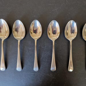 May include: A set of six silver-toned metal spoons arranged in a horizontal row. Each spoon features a smooth, reflective oval bowl and a plain, slender handle that tapers slightly toward the end, showcasing a polished metallic finish throughout.
