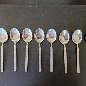 Könnte beinhalten: Sieben silberne Löffel mit ovalen Laffen und dekorativen Griffen liegen auf einer dunklen Oberfläche. Die Griffe weisen ein geätztes Design auf. Die Löffel sind reflektierend, was auf ein Essgeschirr-Set hindeutet.